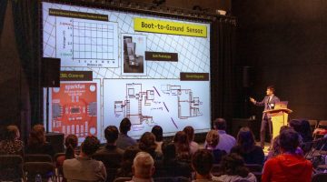 Colby Ting stands at a podium on the right side of the image. The audience is seated facing him, along the bottom of the frame. A large projection screen displays Colby's presentation slide, which reads "Boot-to-Ground Sensor" in a yellow box in the upper left. The slide also includes a chart, a photograph of a boot prototype, and diagrams of a censor and its connections.