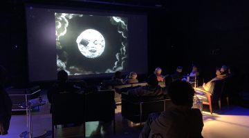 Guests at the IMRC Center are seated in the APPE Space, viewing an early film on the projection screen. The room is darkened, and a small lamp illuminates the right side of the image.