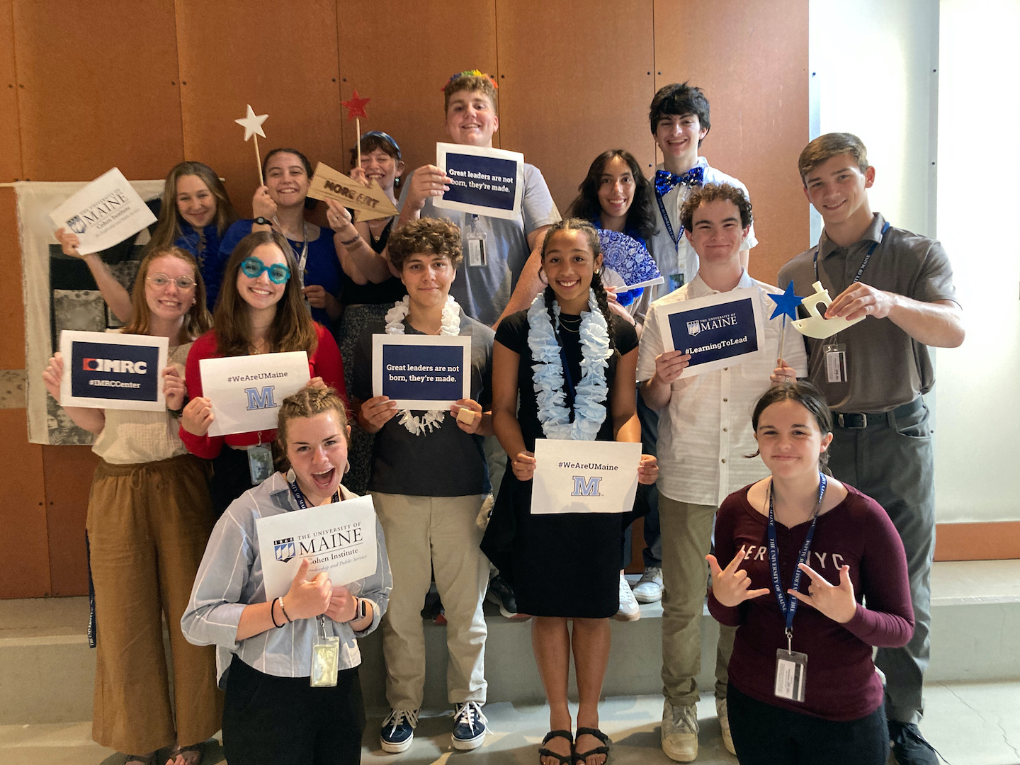 A group of students in professional clothing face the camera. They are holding up signs with slogans and "photo booth"-style props. Some wear oversized sunglasses or leis of artificial flowers.