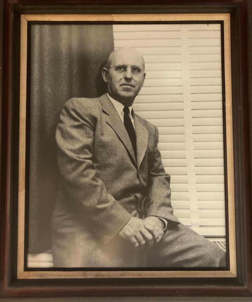 Stewart is seen sitting on a window ledge with a dark curtain and white blinds behind him. He wears a medium-toned suit and dark tie, as well as wire-rimmed glasses, and has a serious but pleasant facial expression. The black and white portrait is framed in dark wood with gold trim.