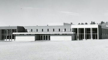 A greyscale photo of Stewart Dining Commons as it appeared after its rededication. A large, close-cut grass lawn spreads out in front of the building. A lower roof covers the front entrance doors, while two large two-story window sections can be seen to either side. A second floor over the main entrance can be seen, with a brick facade and eight smaller windows.